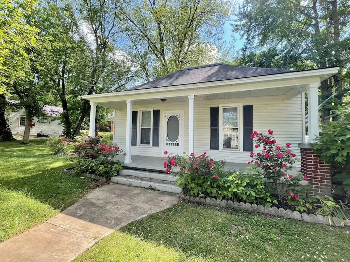 The image features the charming front porch of a newly refurbished yellow cottage, framed by colorful flowering bushes and a welcoming front door. Soft evening sunlight bathes the porch, where comfortable seating invites guests to relax and unwind. Holiday decorations add a festive touch, enhancing the cottage's cozy and inviting atmosphere.
