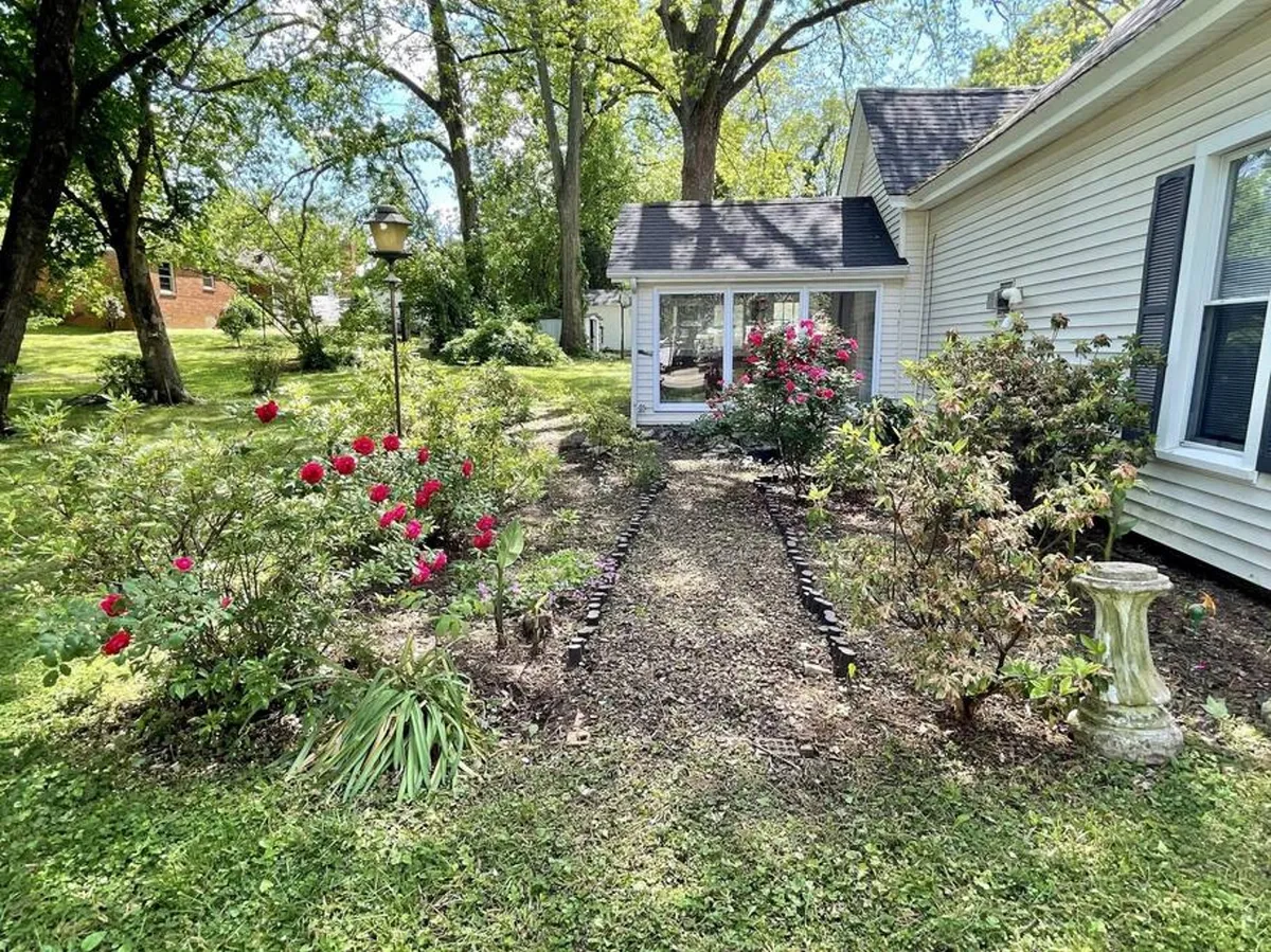 An inviting view of the cottage exterior with flowering bushes, a pathway, and a windowed room visible, suggesting an easy transition from outdoor adventures back to the home. The porch area is subtly visible, hinting at a welcoming space for post-excursion gatherings or unwinding in nature.