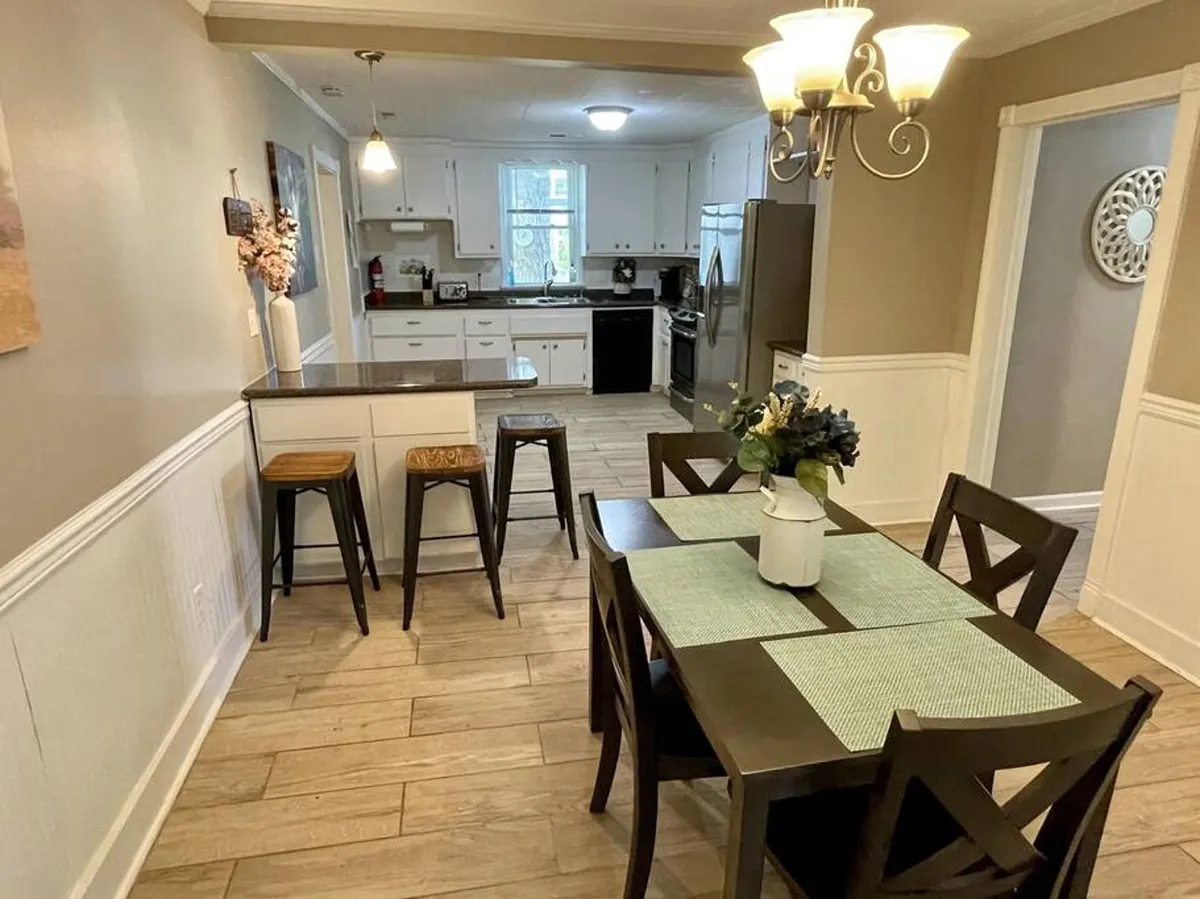 A bright and welcoming dining area connected to a modern kitchen, featuring a spacious dining table, kitchen counter with stools, and a stylish hanging light fixture. The scene is set for family meals, entertaining guests, or casual dining.