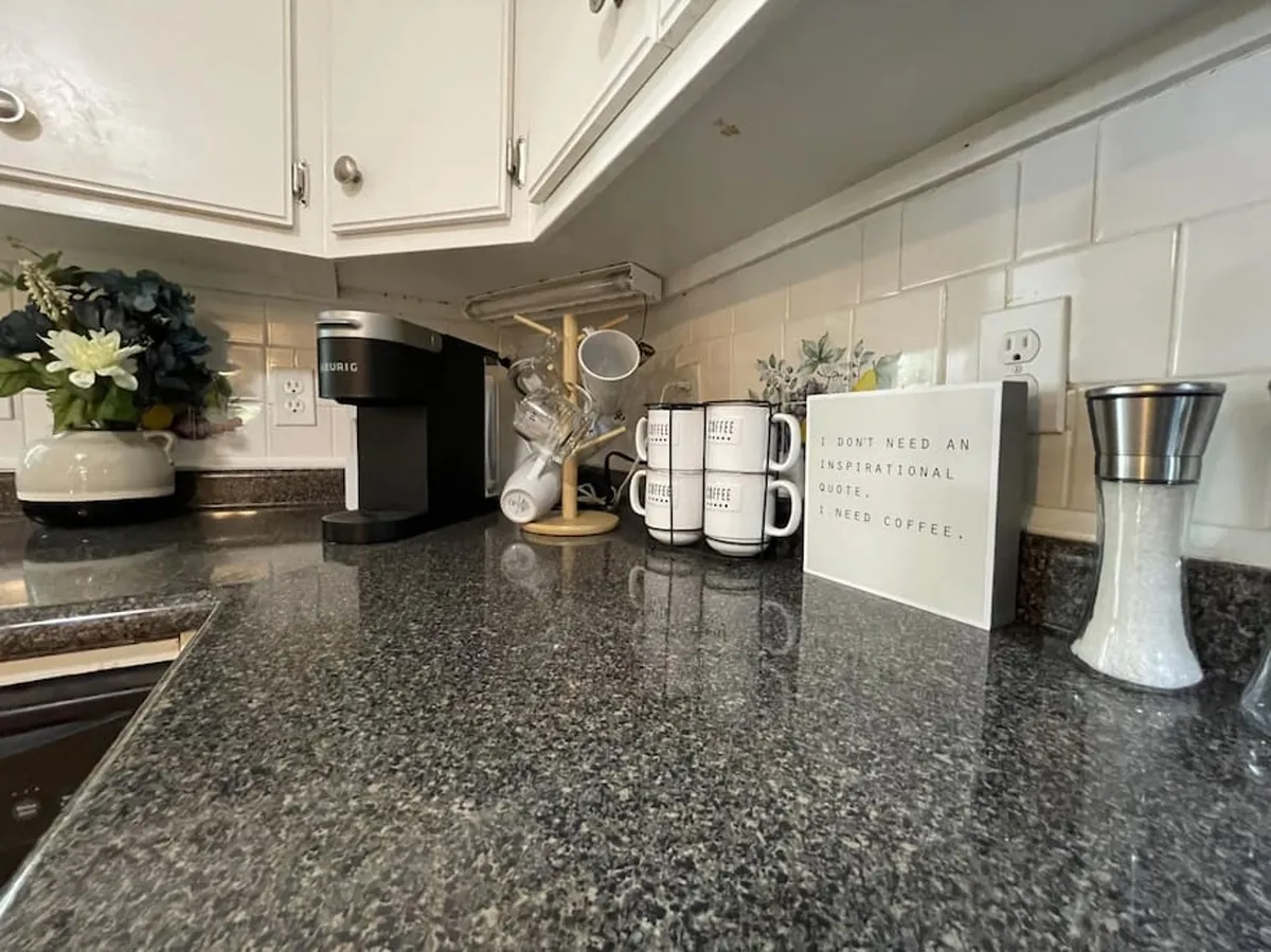 A cozy coffee nook featuring a Keurig coffee maker, mugs with inspirational quotes, a decorative plant arrangement, and a welcoming sign. The setup suggests leisurely mornings and a warm, inviting atmosphere for couples.