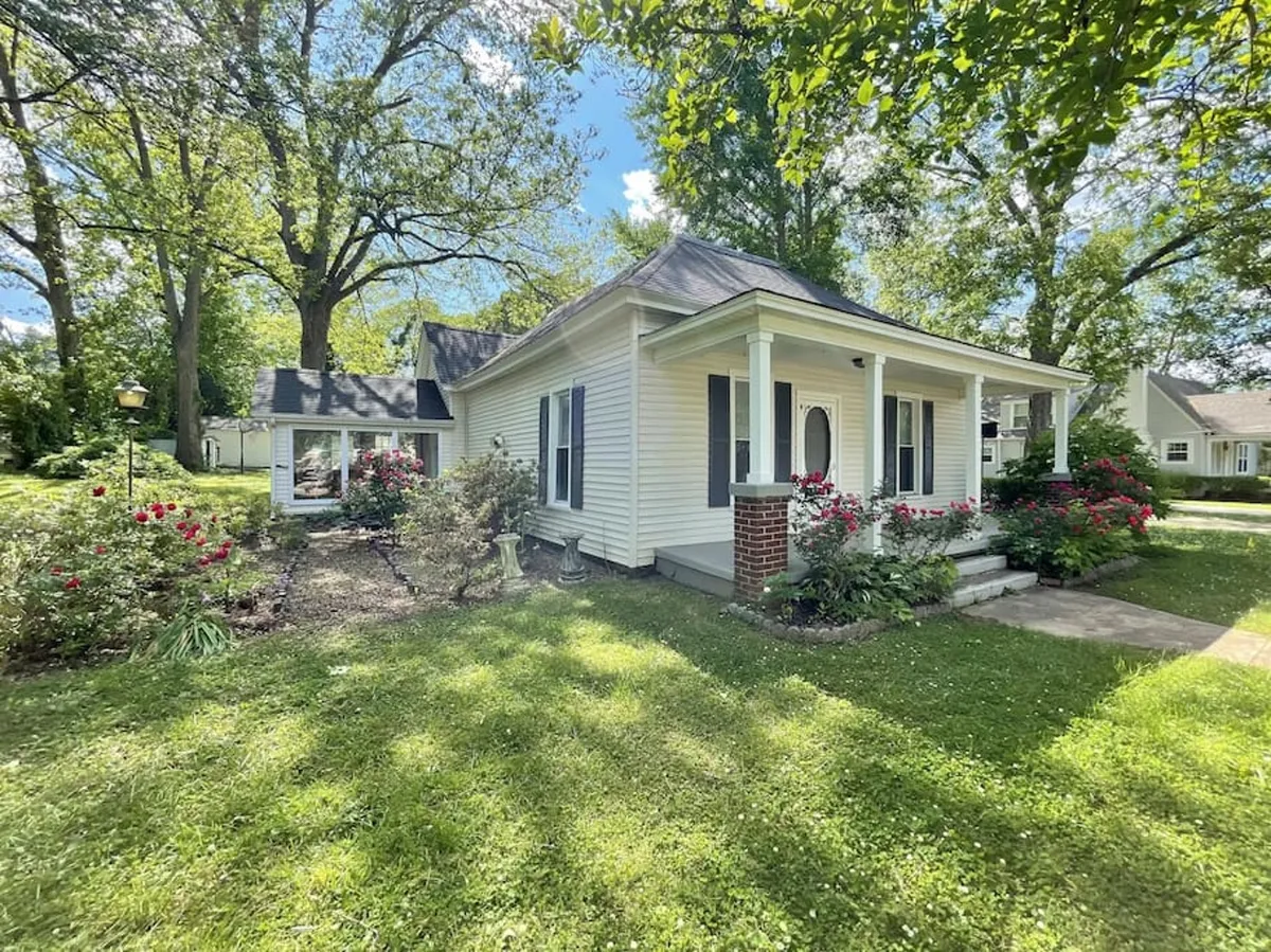 A welcoming exterior view of the cottage's front porch framed by vibrant garden flowers, a lush green lawn, and mature trees. The porch features comfortable seating, inviting guests to pause and relax while enjoying the tranquil surroundings.