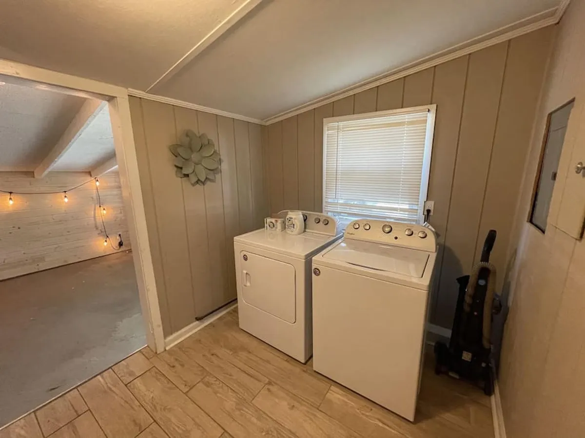 A bright, practical laundry room with a washing machine and dryer beneath a window, featuring decorative wall art that adds a welcoming, homey feel. There is ample functional floor and surface space for sorting and storing gear such as fishing rods, tennis rackets, or picnic equipment, aligning with the section's emphasis on flexible storage for recreational items.