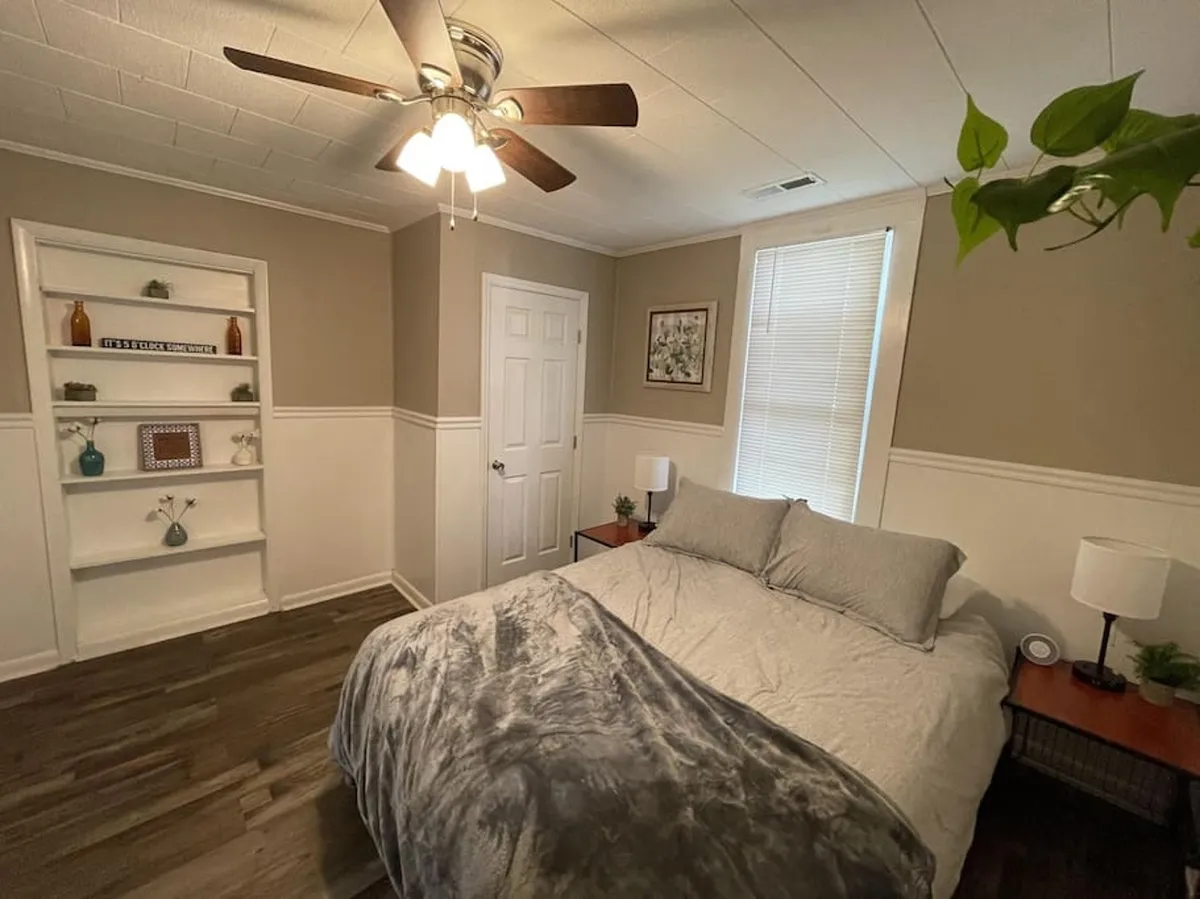A bedroom with a neatly made bed, a bookshelf filled with books, and a window letting in soft, natural light. The room has an inviting atmosphere, ideal for reading or relaxing, and is styled with simple, comfortable furnishings.